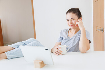 Smiling woman relaxing at home with feet on the table, holding a coffee cup and looking at a tablet. Leisure time, online entertainment, digital lifestyle and home relaxation. © soya