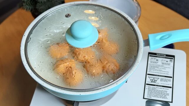 A close up of a hand placing a covered blue frying pan onto a white portable stove. Condensation builds on the glass lid as orange food items steam inside on a warm wooden kitchen table.