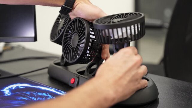 Close-up of male hands adjusting a black triple-head portable cooling fan on a dark office desk. A computer monitor and blue gaming mousepad sit nearby in the bright modern workspace.
