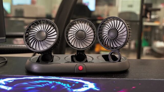 A close-up static shot of a black portable triple-head cooling fan sitting on a modern office desk. The small electronic appliance rests near a colorful mousepad with a blurred corporate background.