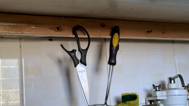 A close-up static shot of kitchen scissors and a metal strainer hanging on a wooden beam. The cooking tools rest against a white tiled wall near a sink and sponge in a domestic indoor kitchen.