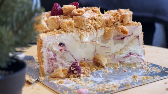 A close-up shot of a sliced raspberry cream pastry cake resting on a silver board. The delicious flaky dessert sits on a wooden table indoors beside a small potted plant in bright lighting.