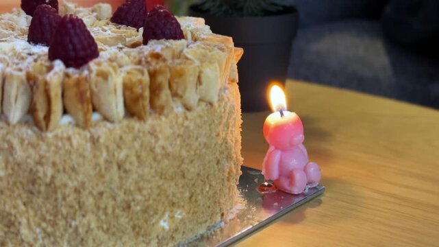 A close-up shot of a lit pink bear candle burning next to a flaky raspberry layer cake. The delicious pastry dessert sits on a silver board over a wooden table indoors with a blurred potted plant.