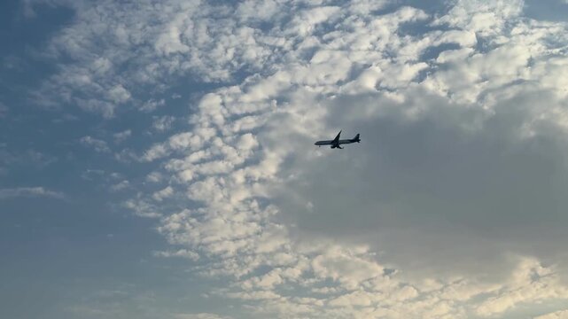A wide shot of a commercial passenger airplane flying through a textured cloudy sky. The dark silhouette of the jet aircraft contrasts against the bright white and gray clouds during the daytime.