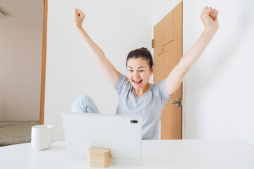 Happy woman using tablet at home, celebrating good news, success or achievement with raised hands and positive emotions. © soya