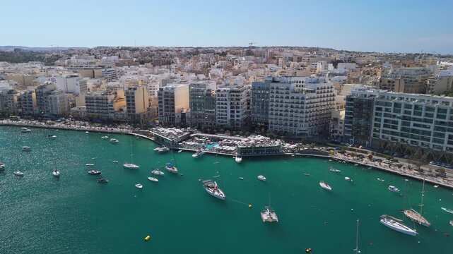 Sliema, Malta waterfront marina with boats, yachts and high-rise apartment buildings along the Mediterranean coastline, featuring promenades and outdoor pools in aerial view.
