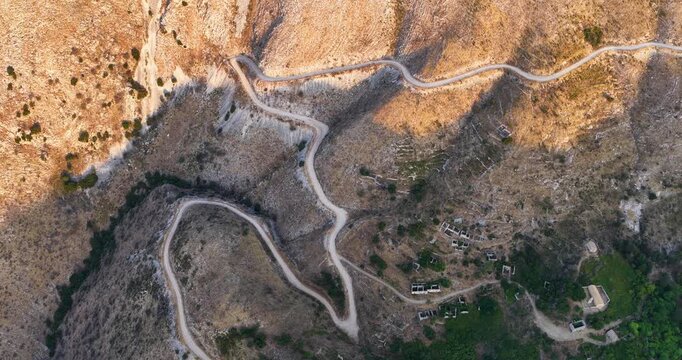 Spectacular top-down drone shot of a serpentine winding road and old abandoned village ruins on the rugged slopes of Mount Pantokrator in Corfu, Greece. Captured at golden hour casting deep shadows.