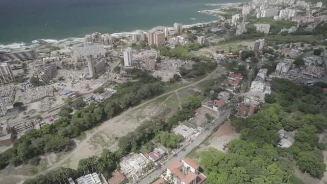 Strong waves crash against shoreline of Caribbean Sea, which borders devastated urban landscape. Ruined structures, collapsed buildings, and massive fields of grey debris and mud cover seaside town