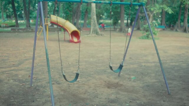 View of empty swings gently hanging in a quiet playground surrounded by trees. Peaceful outdoor recreation area symbolizes childhood memories, leisure, family life and community space
