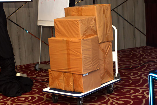 stack of brown wrapped cardboard packages and boxes piled neatly on a white hand trolley cart inside an event room.