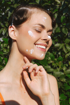 Smiling woman with light skin applying white facial cream outdoors, celebrating natural skin texture. Close-up on sunlit greenery emphasizes skincare, self care, and authentic beauty.