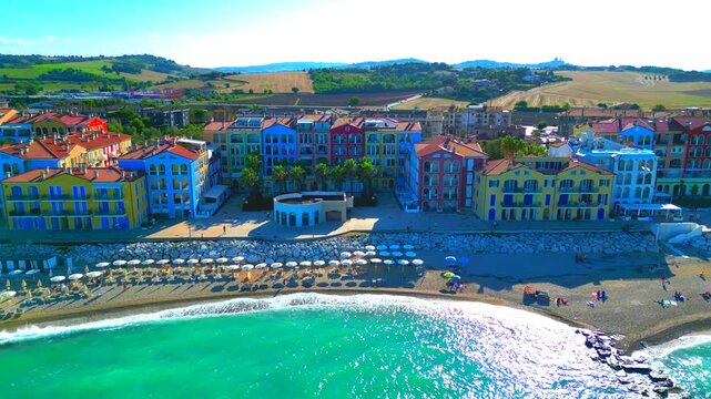 PORTO RECANATI, ITALY - JULY 30, 2025: Aerial pullback pedestal up shot of the beach. Crystalline aquamarine waters, parasols, bathers and pastel-colored buildings right on the seafront, plus hills