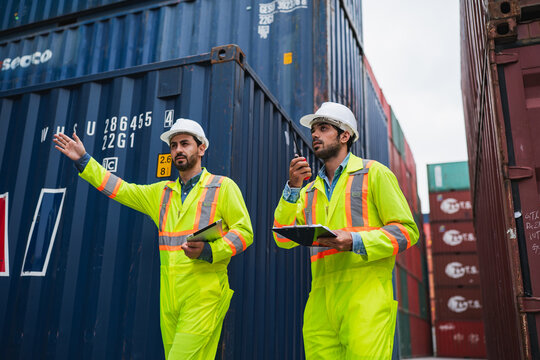 Worker container safety helmet vest cargo shipping yard clipboard radio, two worker inspect shipping container yard with serious expression, wearing safety gear while checking cargo logistics
