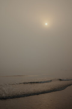 Misty beach with gentle ocean waves and soft golden sunlight filtering through the fog, illuminating the sand and sea in a subdued atmospheric coastal scene.