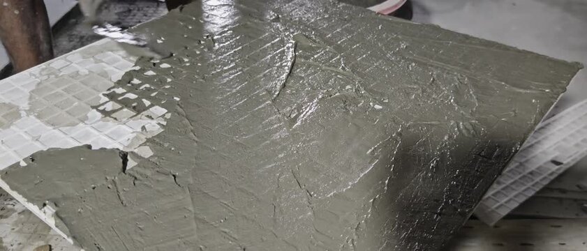 wet cement mortar adhesive spread on back of ceramic tile with trowel during floor installation. Target audience includes construction contractors, tile layers, interior renovation blogs, building