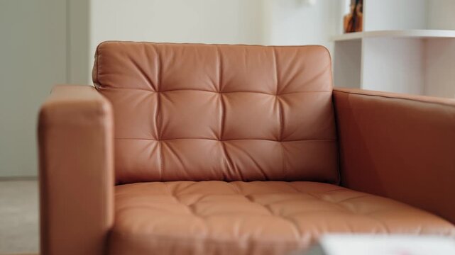 Close-up view of a comfortable brown leather sofa with button tufting in a bright and cozy contemporary living room, featuring a blurred background with a white shelving unit