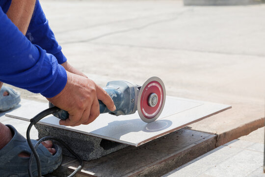 Worker in blue clothing uses an angle grinder to cut a gray tile on a piece of stone. Close-up of tool and process highlights precision and craftsmanship in tile cutting at construction site