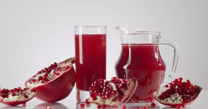 Glass jug and tall glass with delicious ruby red pomegranate juice rotating slowly next to ripe fresh fruit pieces and scattered seeds, all isolated on a clean white background