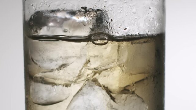 Refreshing amber alcoholic beverage splashing over ice cubes in a cold, condensed glass with water droplets, creating a captivating slow-motion wave effect isolated on a white background