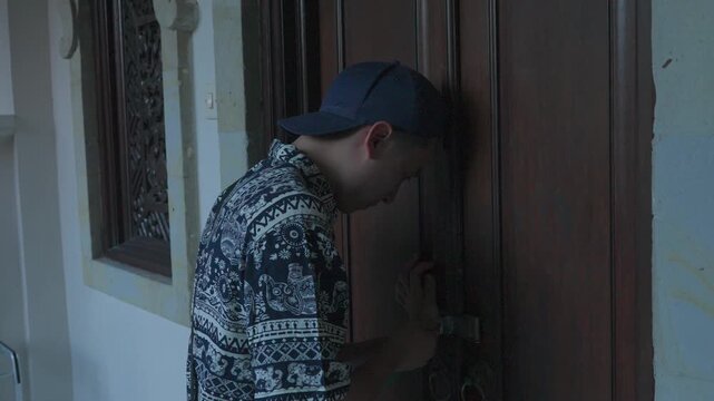 Man bends to work on a wooden door handle. He wears a patterned shirt and cap. His focus suggests careful adjustment or repair. The door has ornate wooden framing