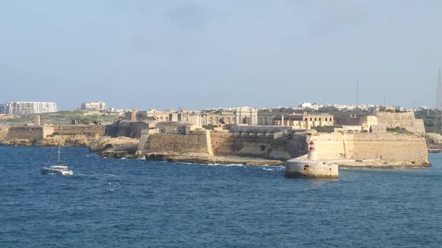 fort ricasoli grand harbour entrance valletta malta sailing catamaran passing lighthouse limestone bastion walls distant apartment towers coastal fortification star fort headland defensive