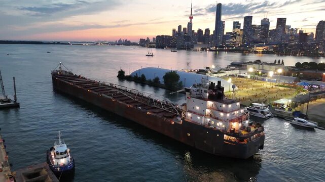 High-angle aerial shot circling the stern, bridge superstructure, and self-unloading boom of the Algoma Buffalo bulk carrier navigating past an illuminated volleyball court plaza toward downtown Toron