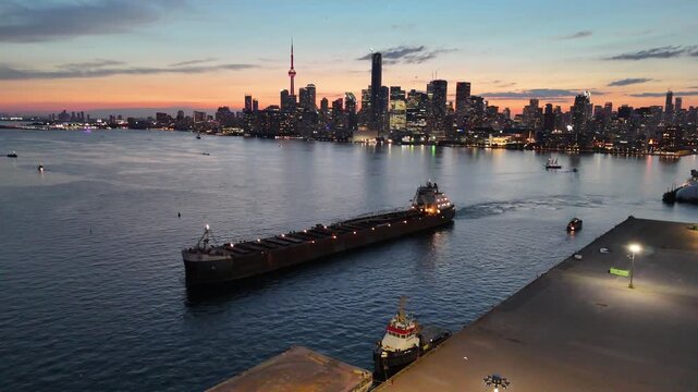 High-angle stationary shot looking down at a large bulk carrier vessel navigating past a concrete industrial pier, revealing the illuminated downtown Toronto skyline and CN Tower at dusk