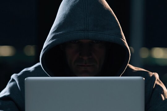 Hacker in a dark hoodie using a laptop to commit cyber crime in the deep darkness