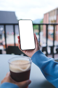 Vertical mockup image of a woman holding and using mobile phone with blank desktop screen while drinking coffee in cafe