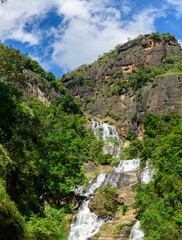 Multi-tiered waterfall cascades down rugged rocky cliffs surrounded by lush green forest in the highlands of Nuwara Eliya, Sri Lanka, beneath a bright blue sky © nilanka