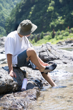 川辺で足を水につけて景色を眺める休憩する夏のアウトドアやレジャーや川遊びのお出かけのイメージ　顔無しの後姿