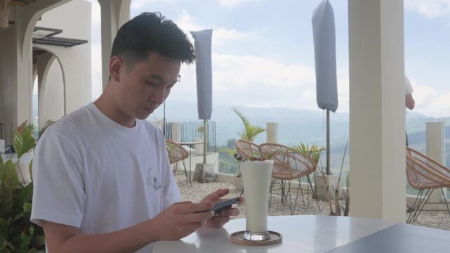 Young man sits calmly at outdoor cafe table. He looks down at his phone with focused attention. Tall drink rests beside him on a small coaster. Soft light fills the scene through open archways