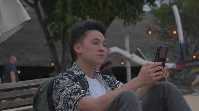 Young man sits quietly on a bench. He holds a phone, looking at its screen. Warm lights glow softly behind him. He wears a patterned shirt and carries a backpack