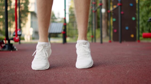 Sportsman preparation before a workout. Man in white running sneakers doing light warm up jumps on an outdoor sports ground. Athlete doing jumping exercises on an outside gym.