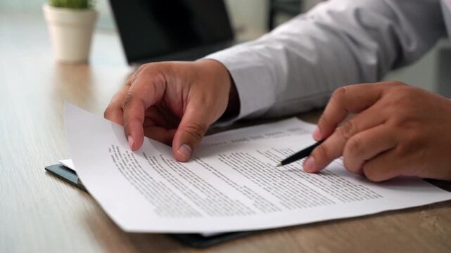 businessman signing over contract papers on clipboard at wooden desk spreadsheet, laptop, glasses, preparing agreement signing, document review, and business paperwork workflow in daylight.