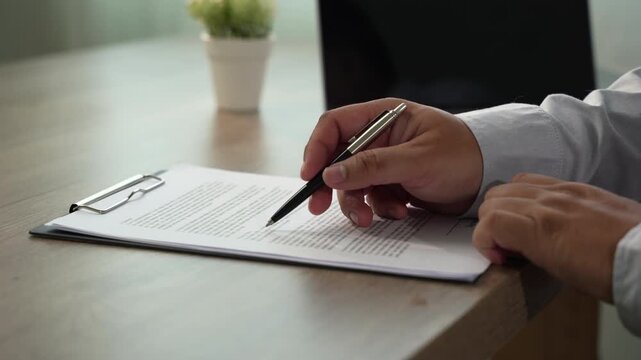 businessman signing over contract papers on clipboard at wooden desk spreadsheet, laptop, glasses, preparing agreement signing, document review, and business paperwork workflow in daylight.