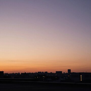 city skyline silhouette at dusk seen from a rooftop, large empty sky with orange and purple gradient, a few distant buildings with warm glowing lights