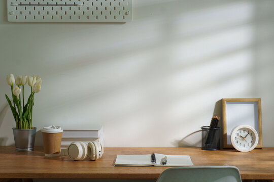 Minimal wooden desk workspace with calculator, coffee mug, books, headphones, plant, pencils, and clock against a bright wall with copy space.