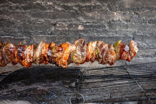 Traditional Greek kontosouvli pork meat chunks marinated with spices and bell peppers roasting slowly on a horizontal metal spit over hot charcoal embers capturing smoke and sizzling details.
