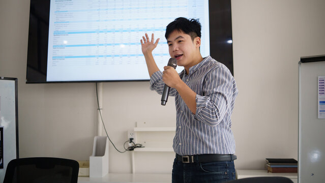 Business professional giving a presentation with a microphone while explaining spreadsheet data and financial information displayed on a large screen.