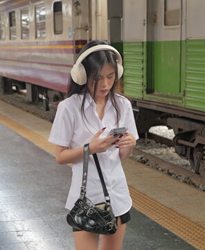 A young Asian woman wearing a university student uniform and over-ear headphones stands on a train platform looking down at her smartphone with a black handbag slung over her arm
