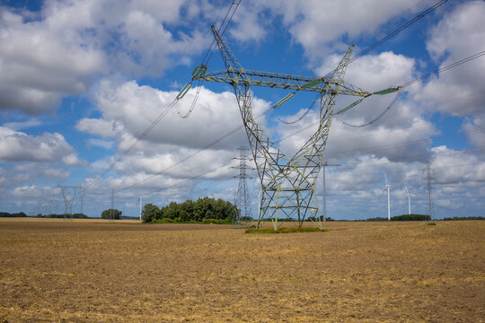 High Voltage Electricity Pylon across Harvested Field with Wind Farm and Summer Clouds. Dramatic rural landscape centered on a large high-voltage electricity transmission pylon standing across a harve
