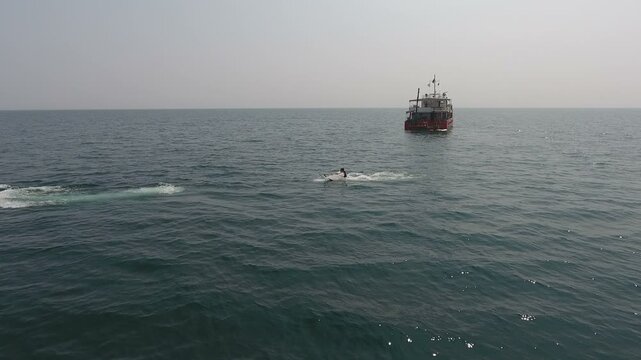 Aerial view of a red inflatable boat towing a rider across open sea, a support vessel anchored in the hazy distance and a long foam wake across the rippled surface