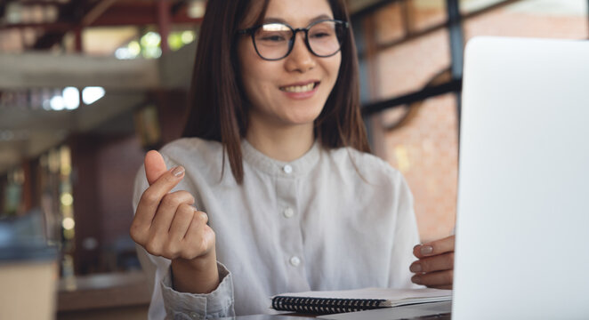 Smiling young asian businesswoman making finger heart gesture on video call, working on laptop in cafe workspace.