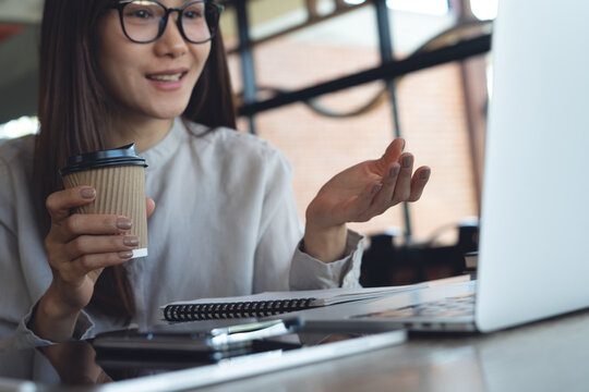 Smiling asian businesswoman holding coffee cup during video call on laptop in cafe, remote work, freelance at work, business casual lifestyle concept.