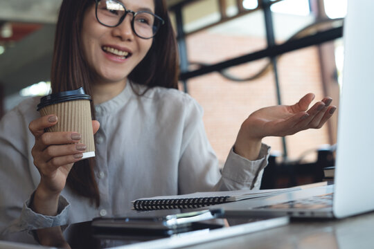 Smiling asian businesswoman holding coffee cup during video call on laptop in cafe, remote work, freelance at work, business casual lifestyle concept.