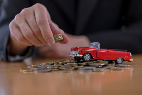 Hand placing coins beside a red toy car on money, representing car insurance, vehicle costs, financial planning, savings, ownership expenses, transportation budget and automotive finance concept.