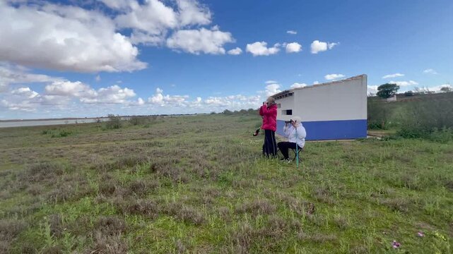 Mother and teenage son birdwatching with binoculars from a nature observation area beside a lagoon during a countryside hike
