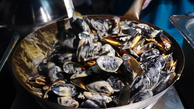 Close-up of large black mussels stirred thoroughly with heavy metal spoon inside hot pan. Street food cooking process, fresh seafood preparation, bustling culinary atmosphere, outdoor dining 4k