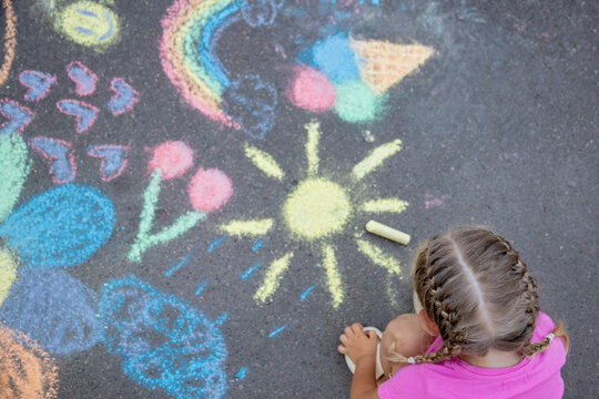 Little girl drawing sun with sidewalk chalk on asphalt, top view